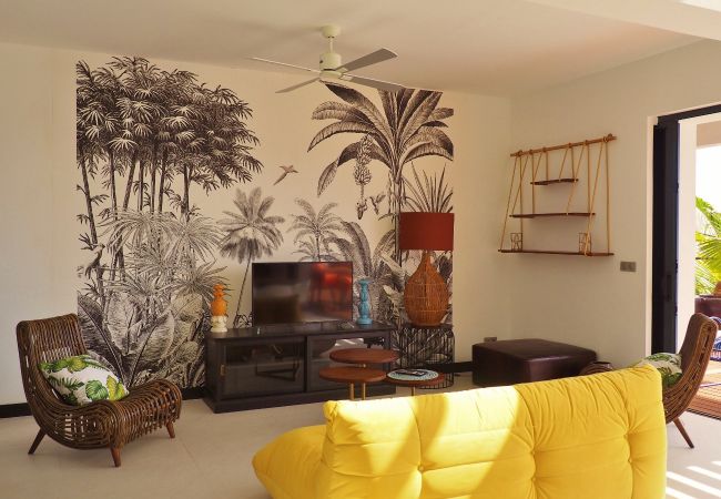 Salon avec canapé jaune, fauteuils en rotin et fresque murale de palmiers – Villa Naézora, villa 10 personnes avec piscine à Saint-Gilles-les-Bains, La Réunion